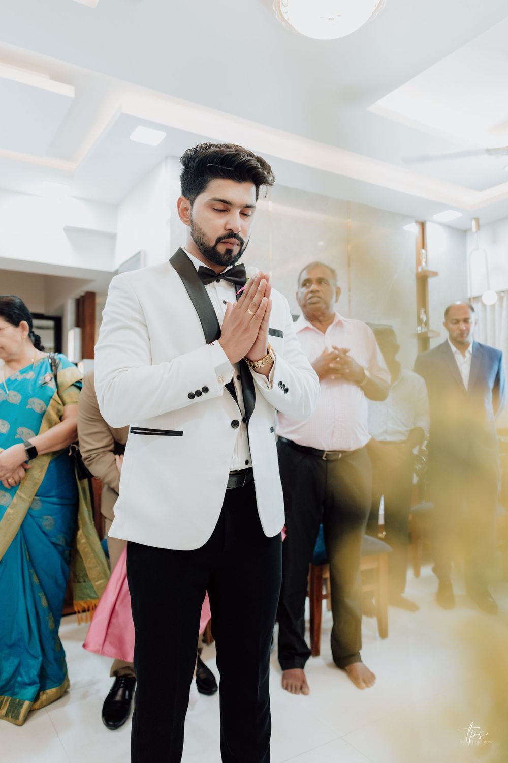 Person in white tuxedo jacket stands in prayer pose during wedding ceremony.