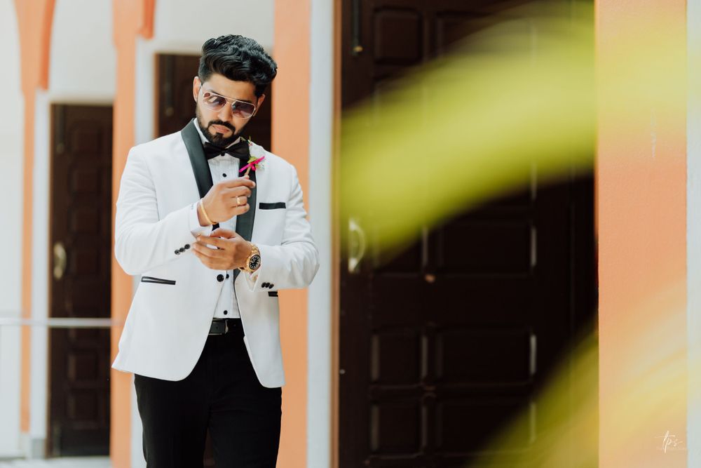Groom in a white tuxedo jacket and black bow tie stands against a peach-colored wall.
