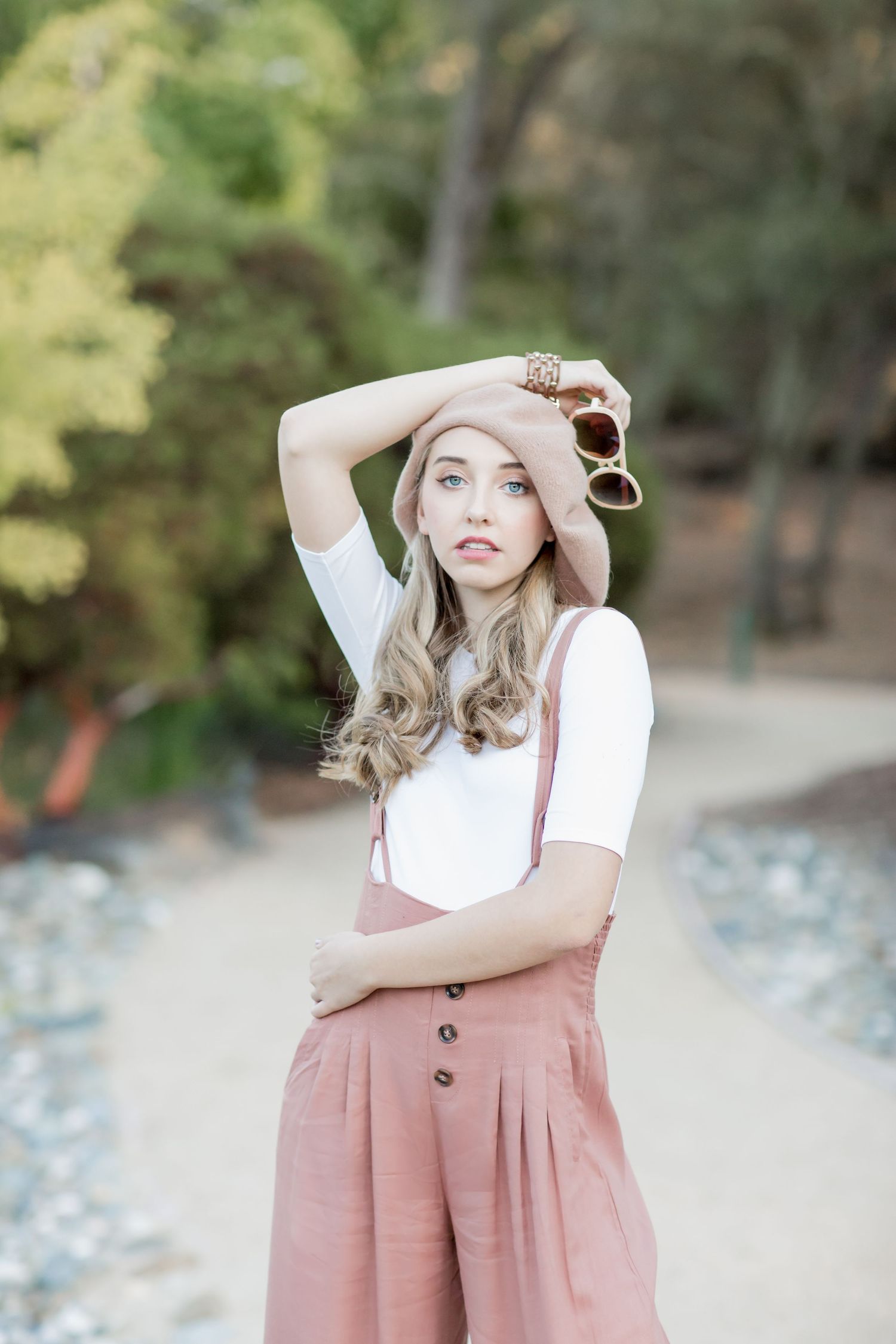 Stylish young woman in pink overalls and hat posing on a scenic outdoor path among trees.