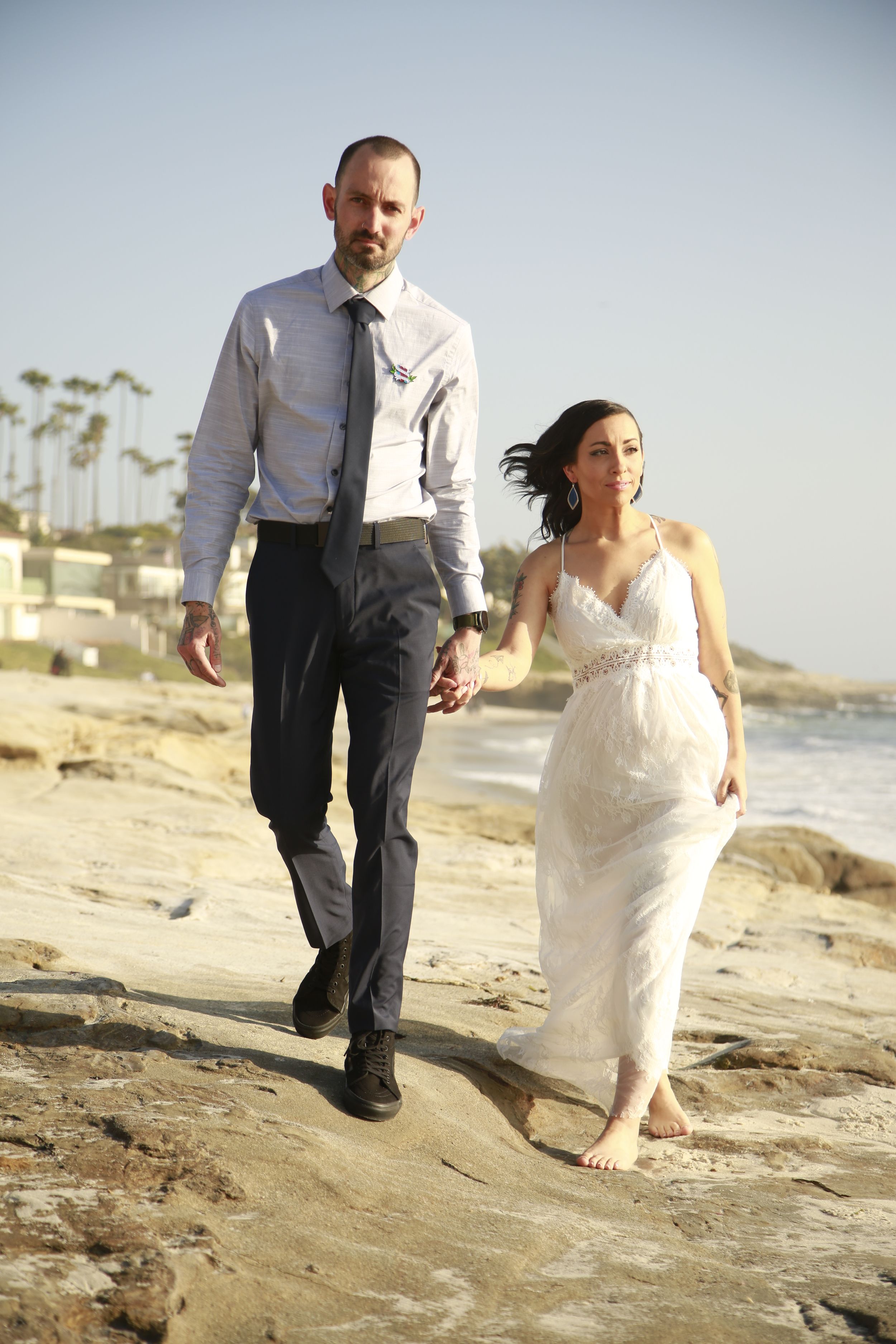 Wedding couple walking along rocky beach shore during sunset photoshoot.