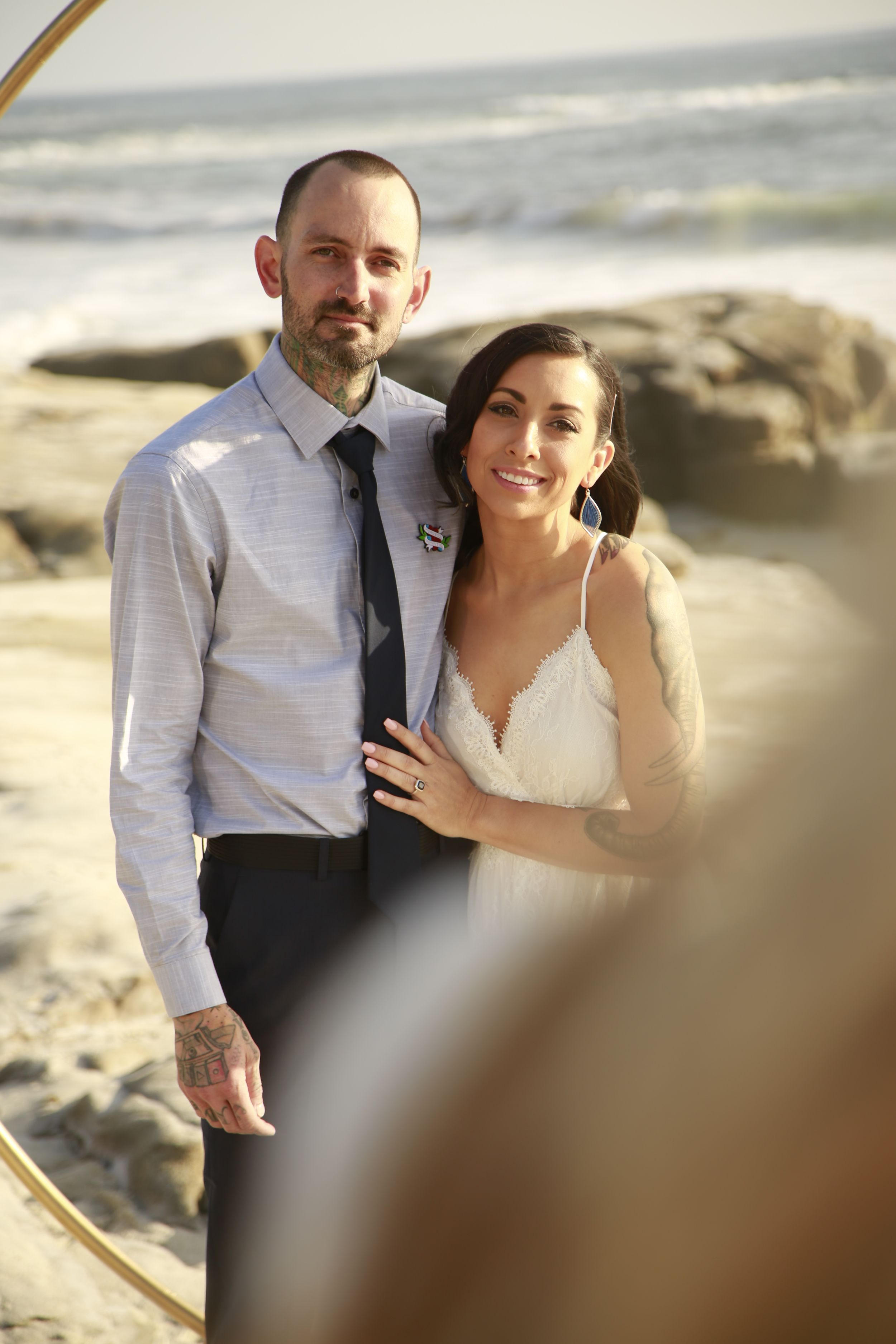 A coastal wedding portrait framed by a circular golden arch at sunset.