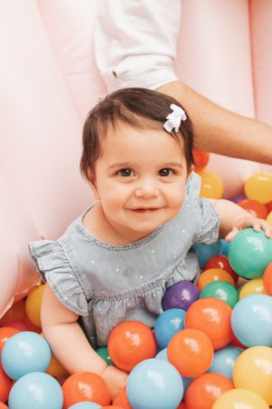 Toddler in blue dress playing in colorful ball pit with joyful expression.