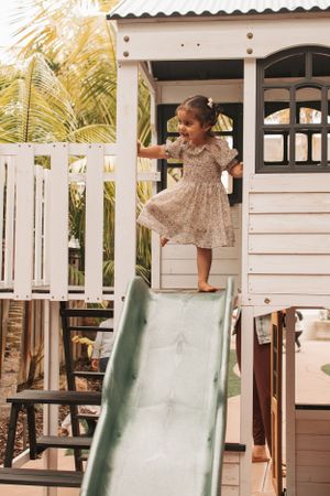 A child in a floral dress plays on a white playhouse with a green slide.