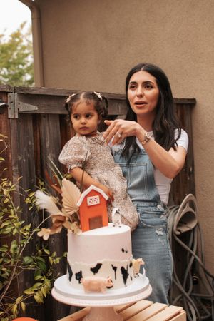 Woman in overalls stands next to white cake decorated with red barn design at outdoor party.