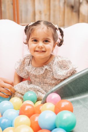 Child in lace dress smiling while playing with colorful balls at party.