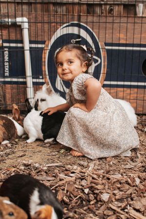 A child wearing a sparkly dress kneels near white rabbits and guinea pigs in a backyard pen.