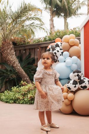 A toddler in a pink dress poses by a balloon arch with soccer ball patterns against a tropical backdrop.