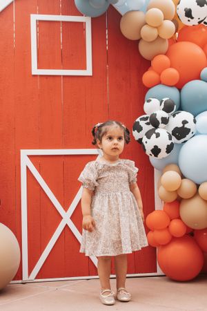 A small child in a floral dress stands in front of a red barn wall decorated with balloons.