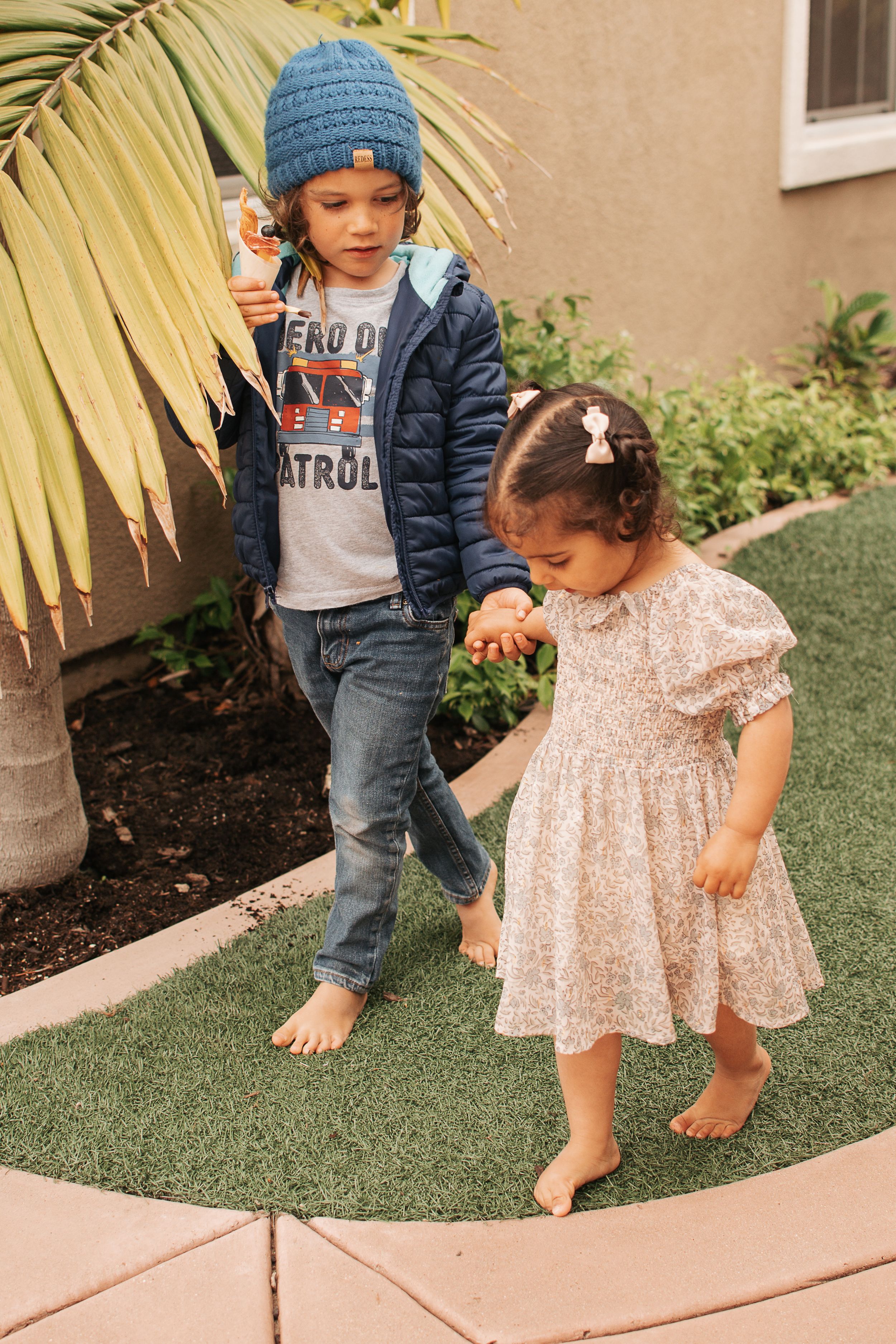 Children playing barefoot on grass near palm trees and building.