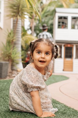 Child in sparkly dress plays on artificial grass with outdoor decor in background.