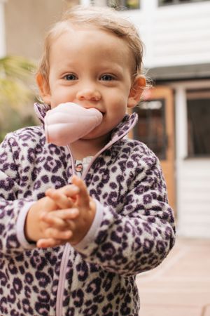 A small child in a leopard print jacket enjoys a pink ice cream cone outdoors.