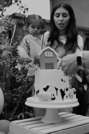 Black and white photo of party scene with decorated cake featuring miniature barn and floral accents.