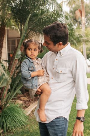 Parent wearing white shirt holds child in denim dress in outdoor garden setting.