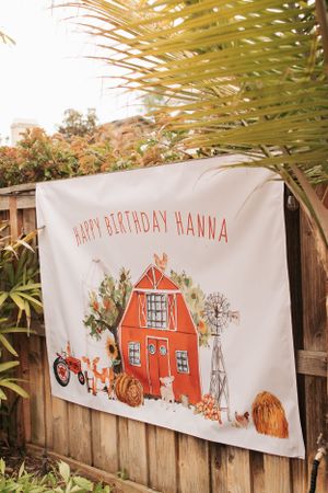 Farm-themed birthday banner with red barn artwork and autumn decorations hanging on wooden fence.