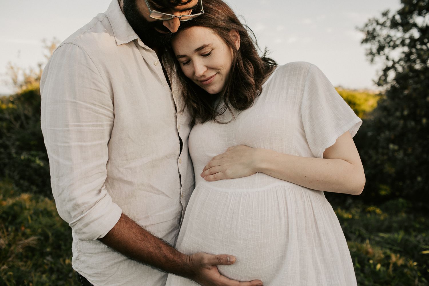 A couple shares a tender embrace at golden hour, gently cradling a pregnant belly in soft natural light.