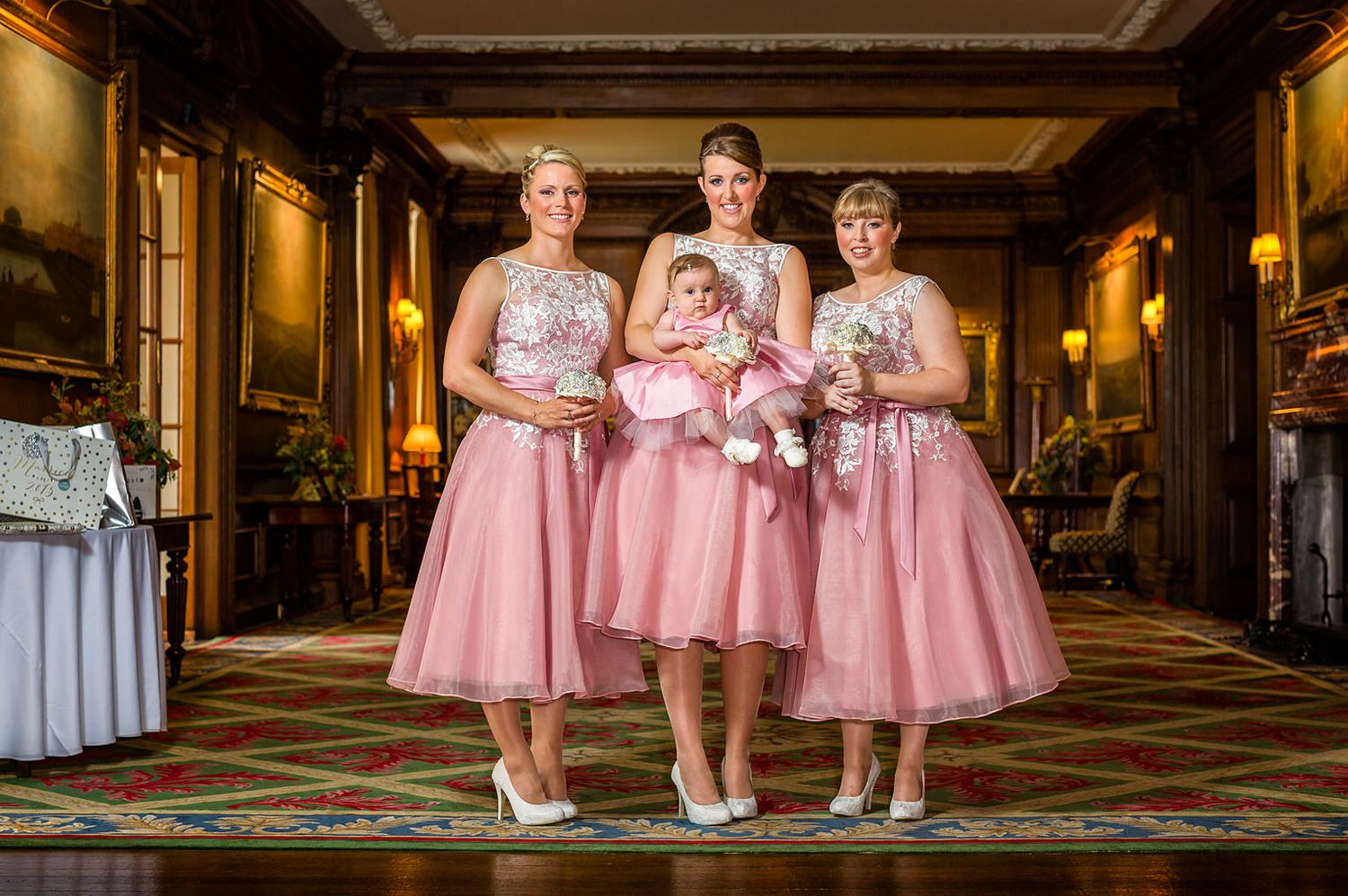 Three bridesmaids wearing pink dresses hold a small baby also in pink indoors at a Knowsley Hall wedding