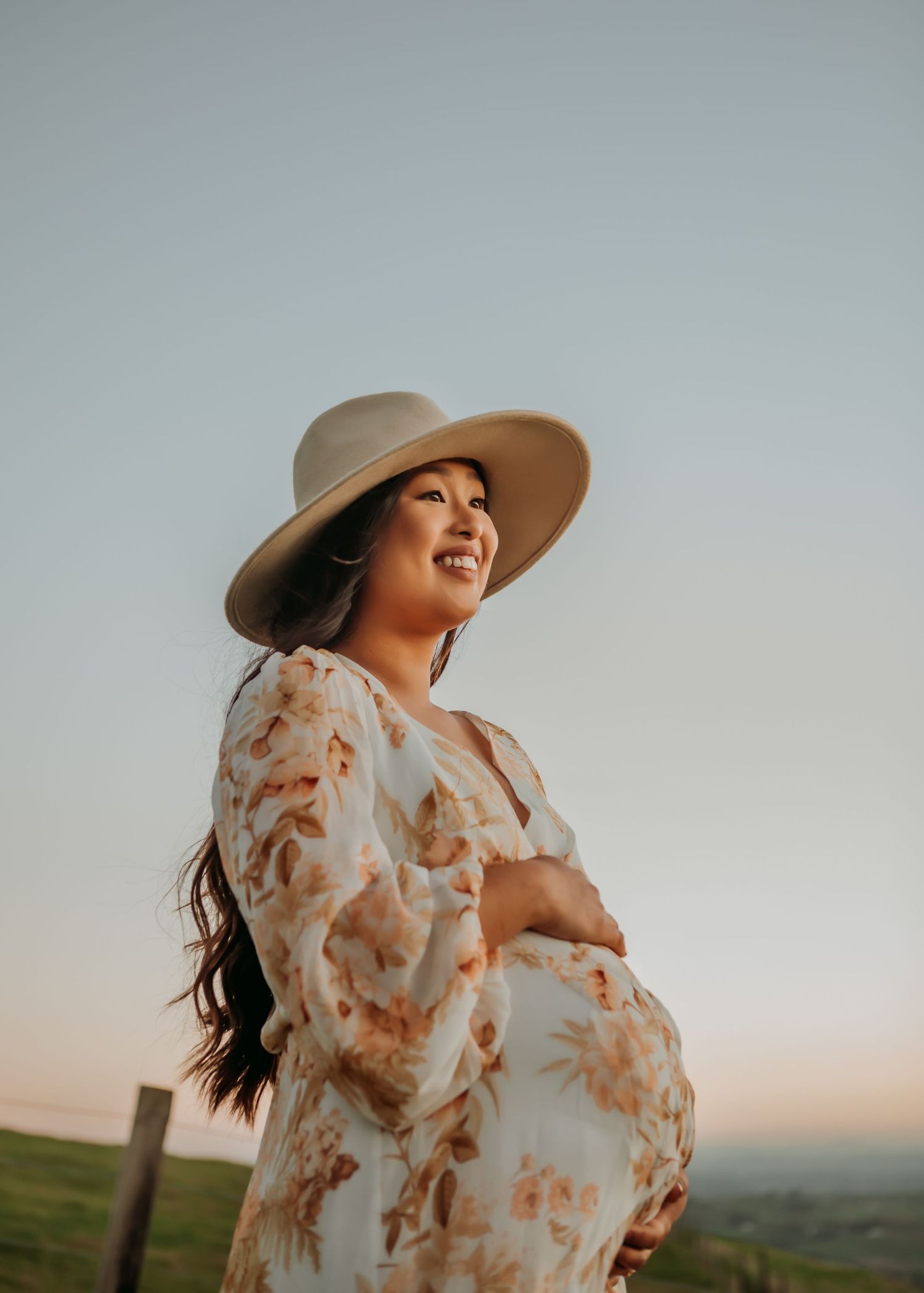 A person in a floral dress and wide brim hat poses in a golden field at sunset.