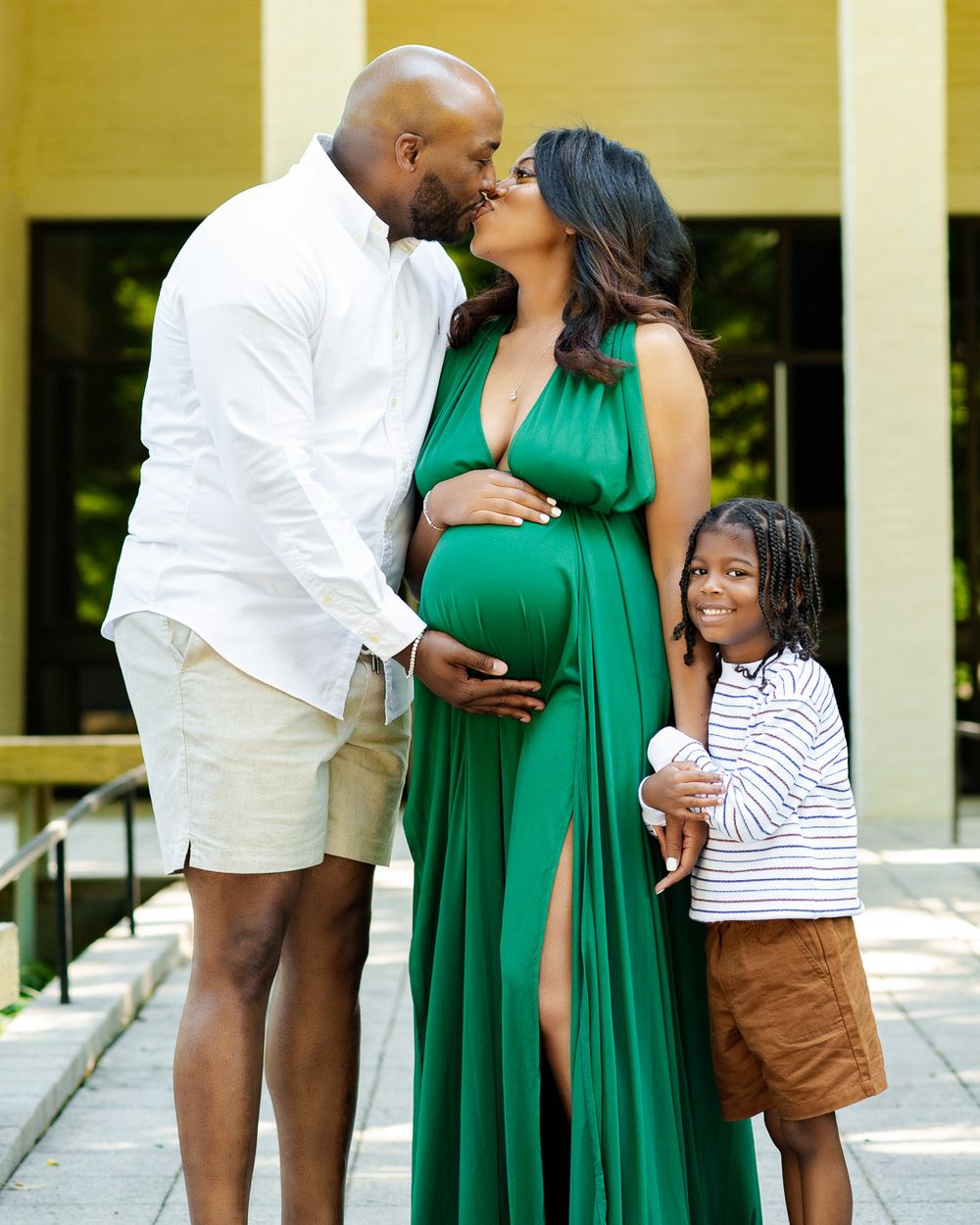 Family in summer attire poses outdoors with expecting mother in flowing green dress.