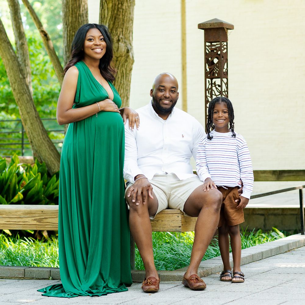 Family sitting on wooden bench outdoors with mother in elegant green maternity gown.
