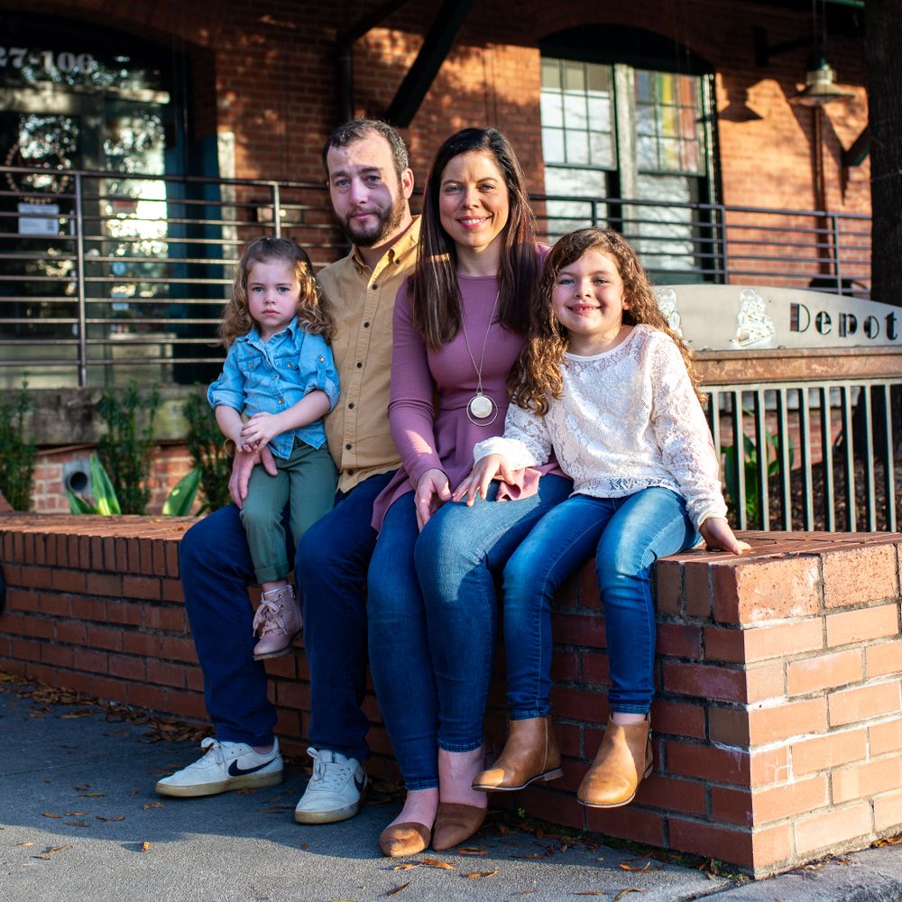 Family of four sitting together on brick ledge outside rustic building.
