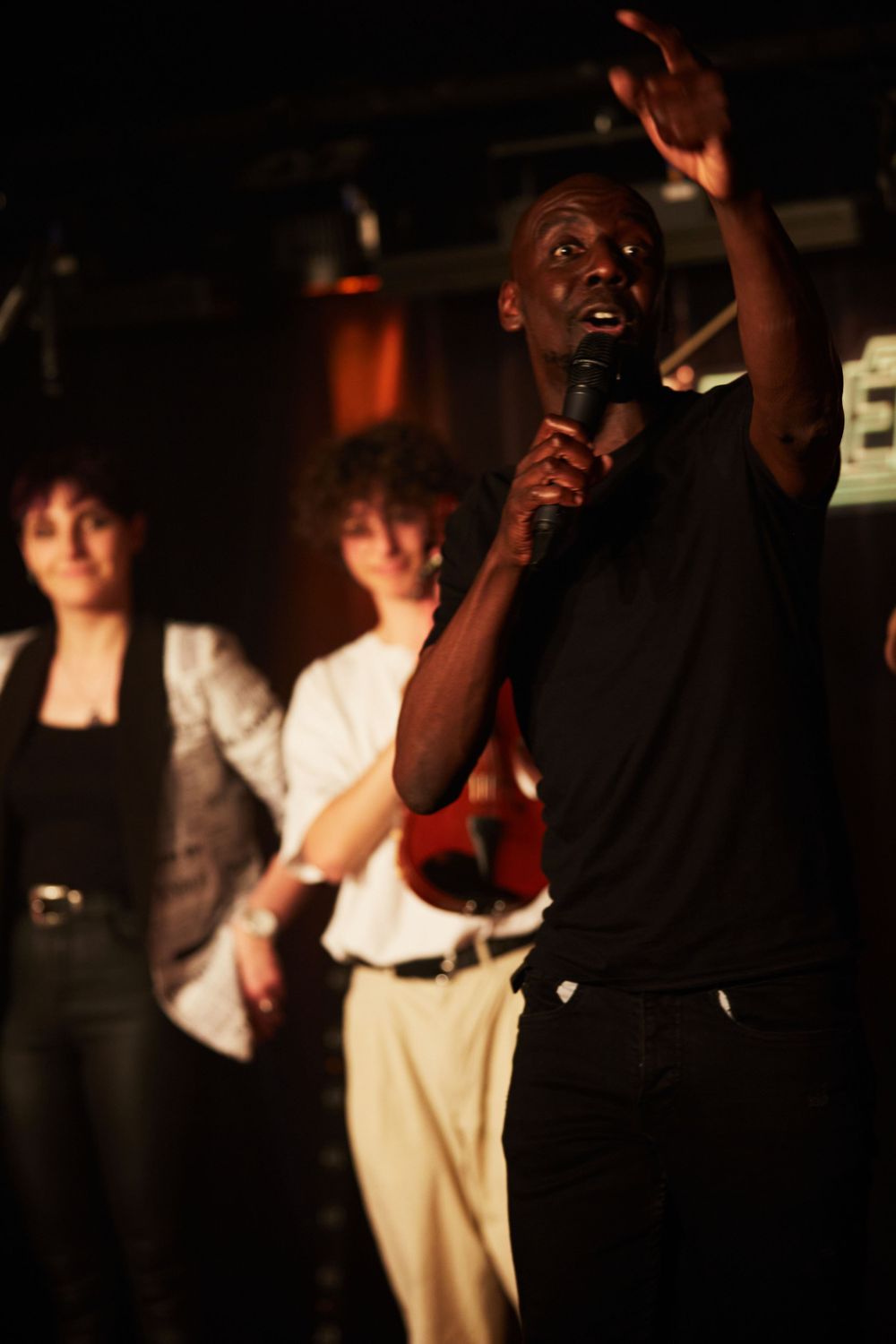 Performer singing into microphone with raised hand while band members play in background.