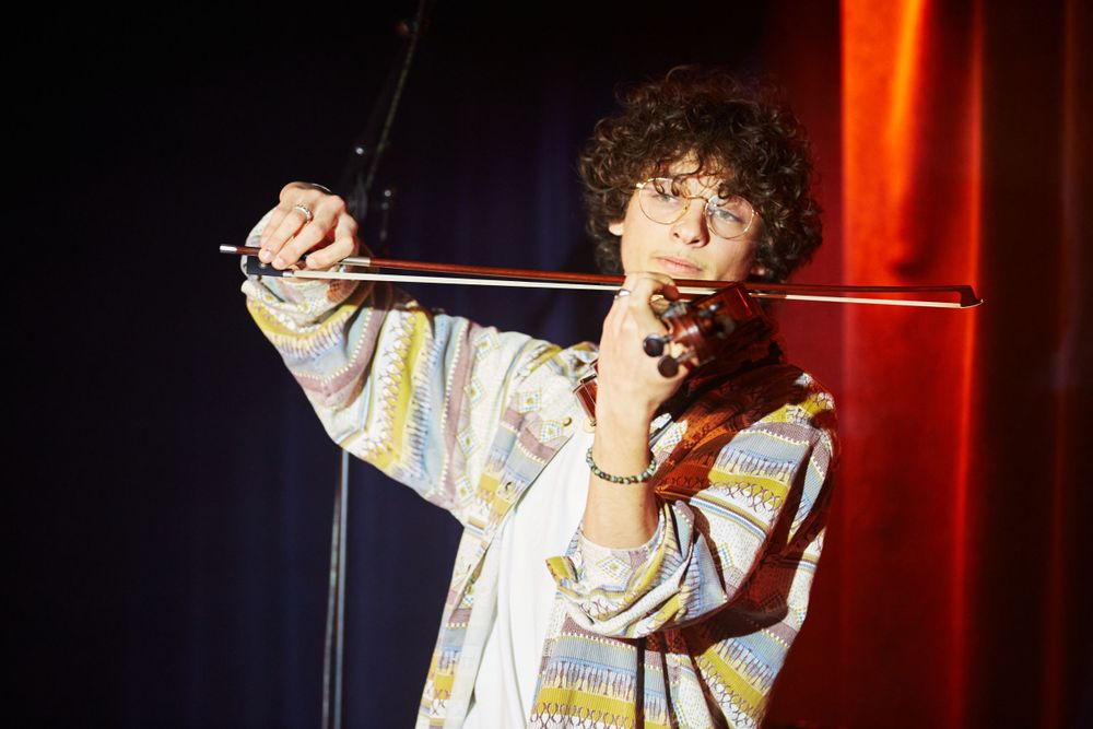 Musician playing violin on stage wearing colorful patterned sweater with dramatic red lighting.