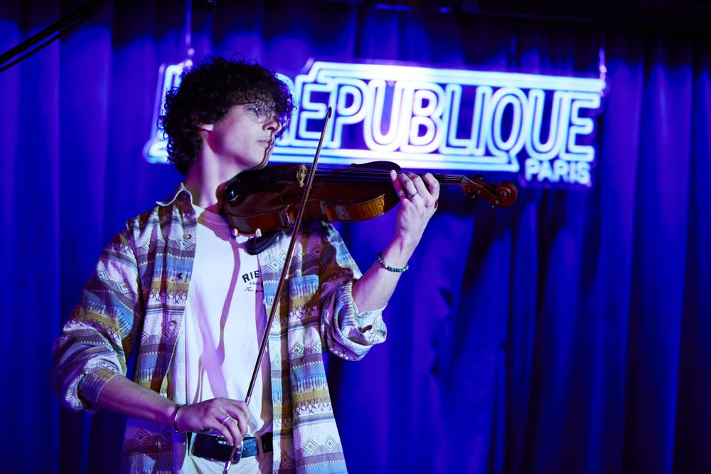 Musician performing with violin under blue neon 'Republique' sign on stage during concert.