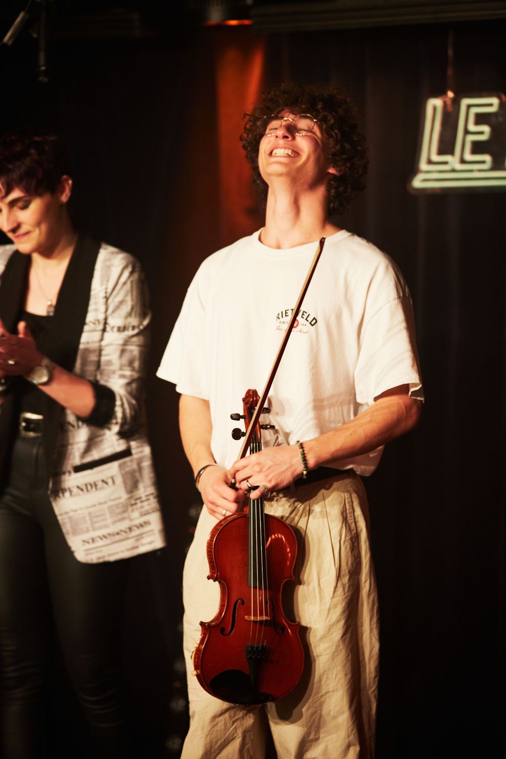 Musician smiling while holding violin during performance on dimly lit stage.