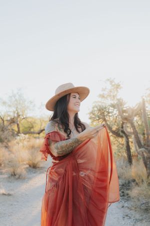Woman in red dress and wide brim hat dances through desert landscape at sunset.