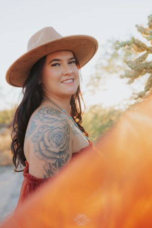 A close-up portrait in warm sunlight showing a rose tattoo and tan hat against desert scenery.
