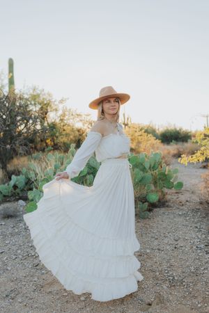 A white prairie dress billows in the desert wind among prickly pear cacti and mountain views.