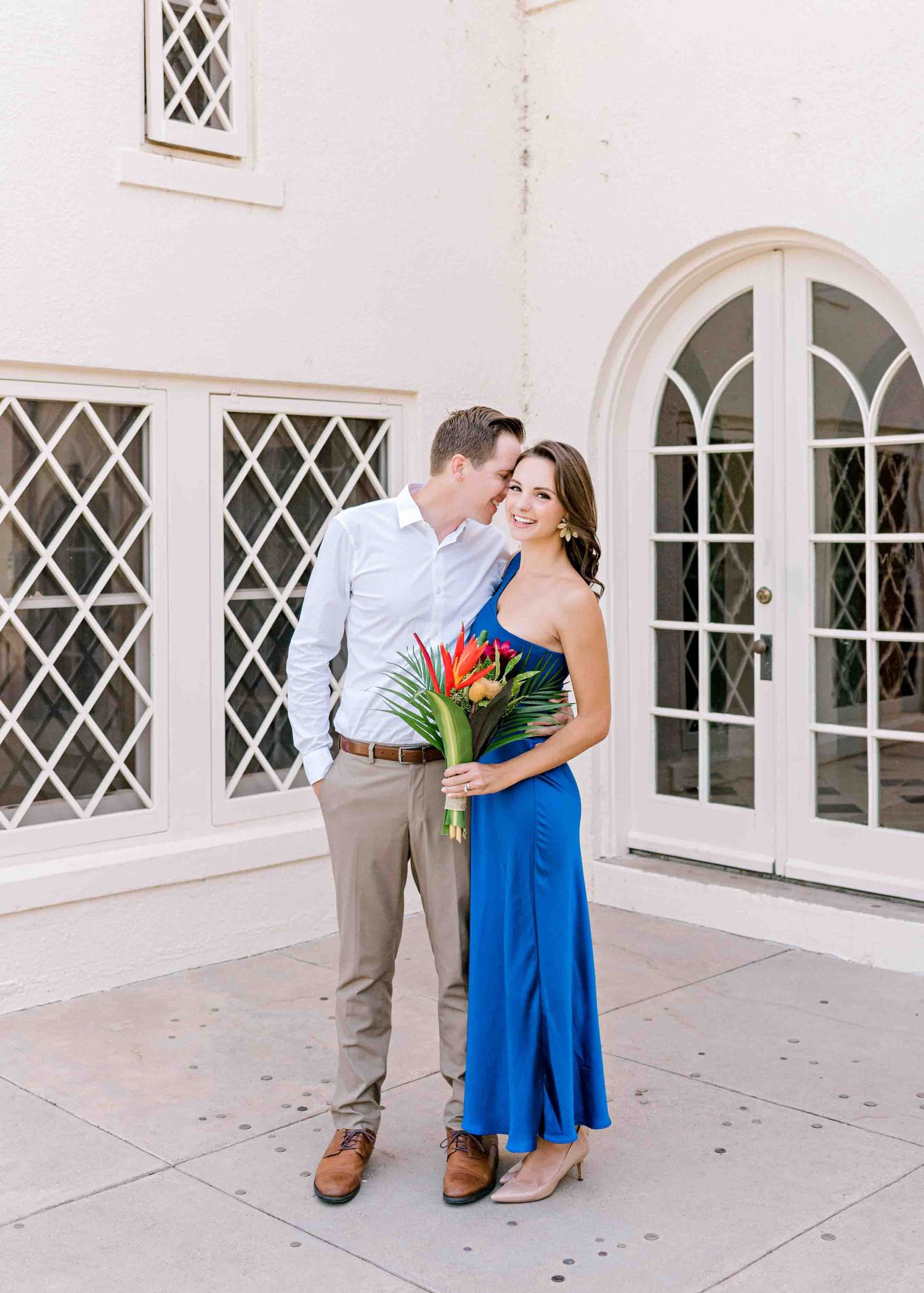 A couple poses outdoors in front of arched windows, with her wearing a blue dress and holding tropical flowers.