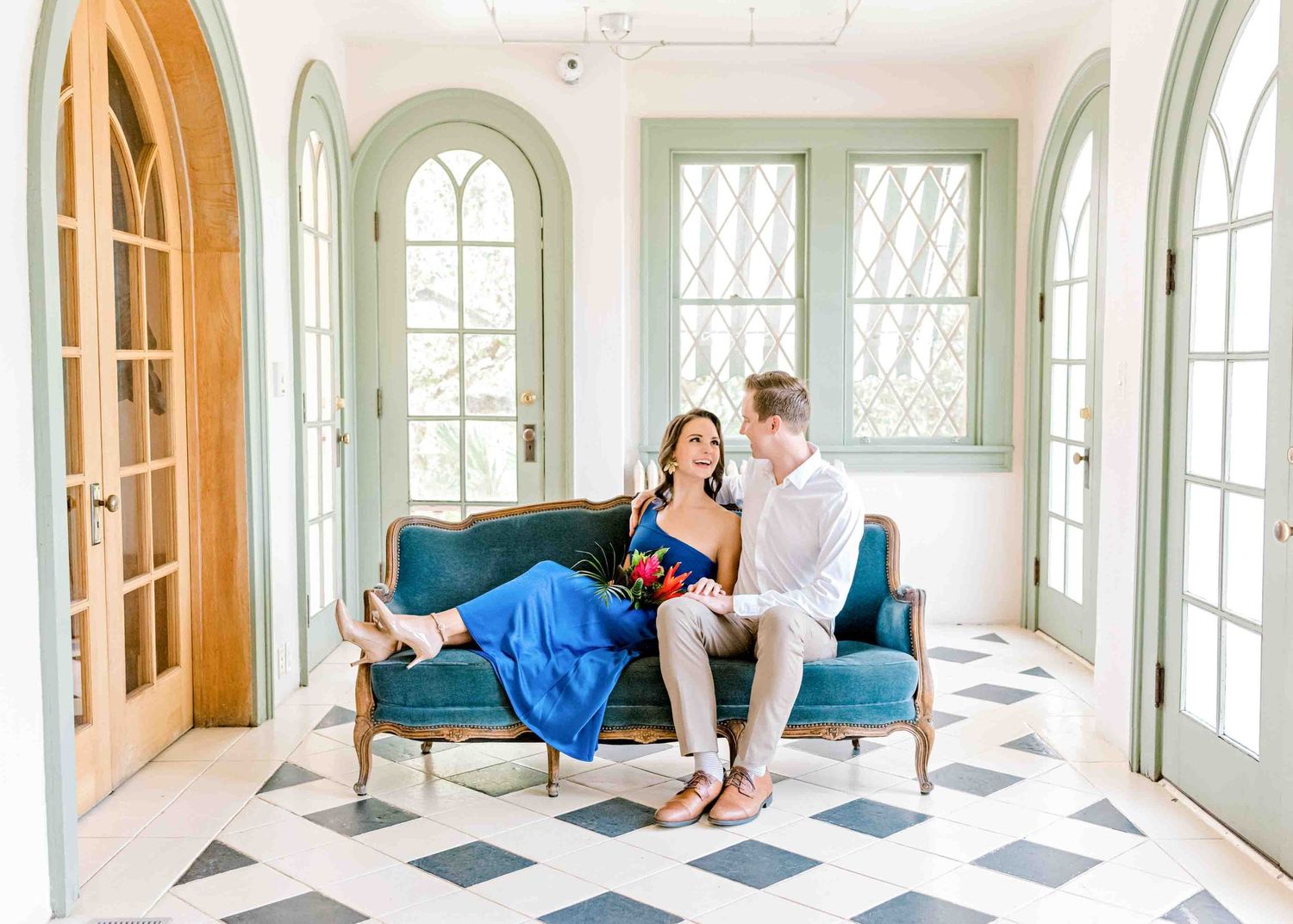 A couple relaxes on blue velvet chairs in a bright sunroom with checkered floors.