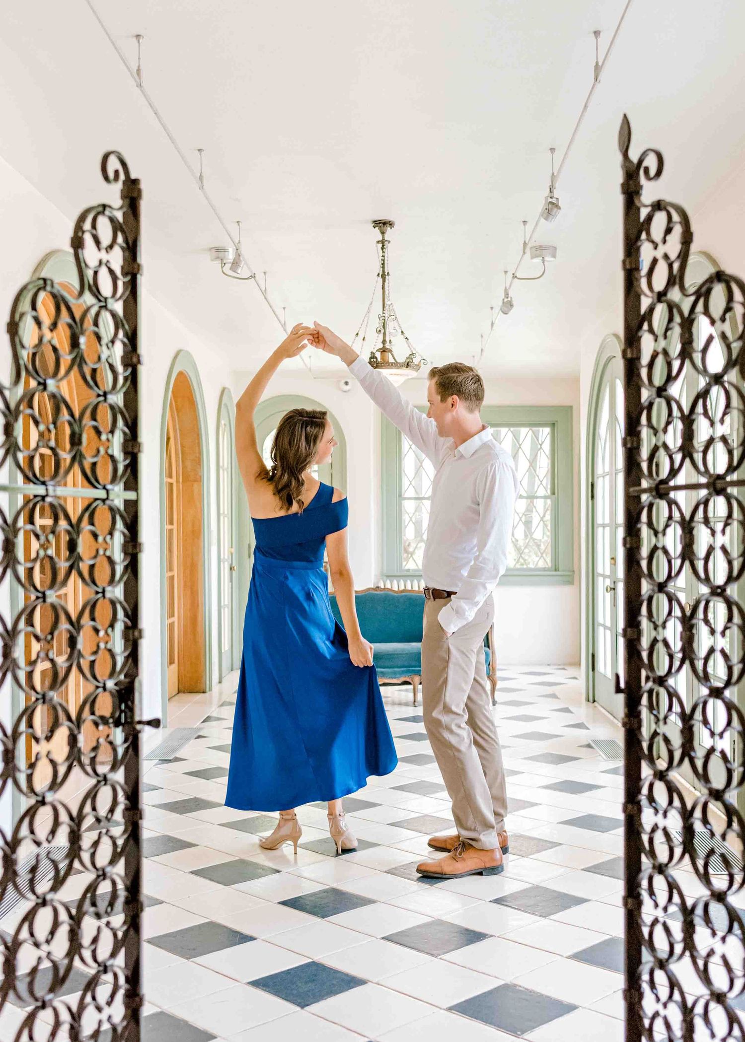 A couple stands between ornate iron gates in a hallway with checkered floors.