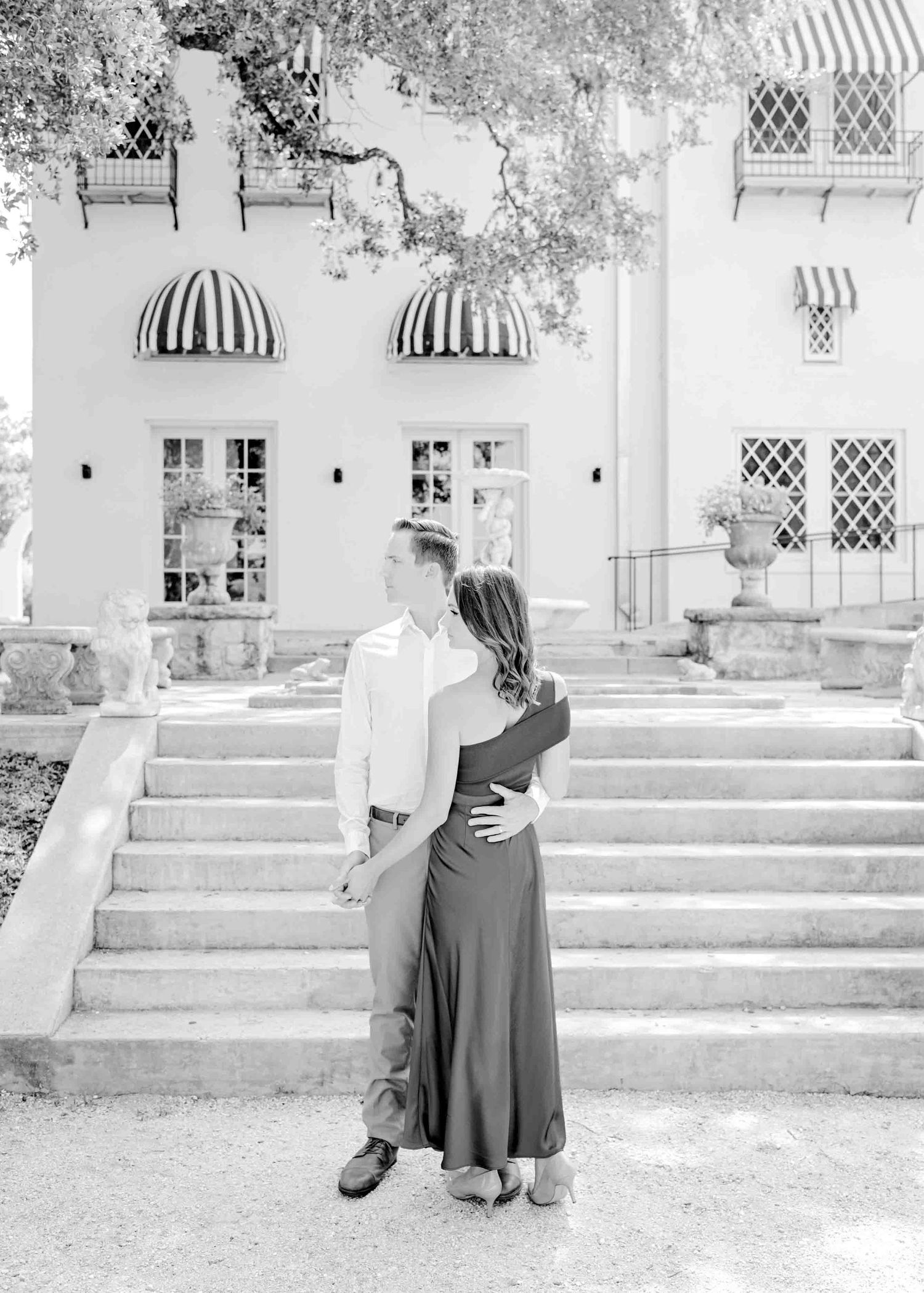 A black and white photo of a couple embracing on elegant stone steps with striped awnings overhead.