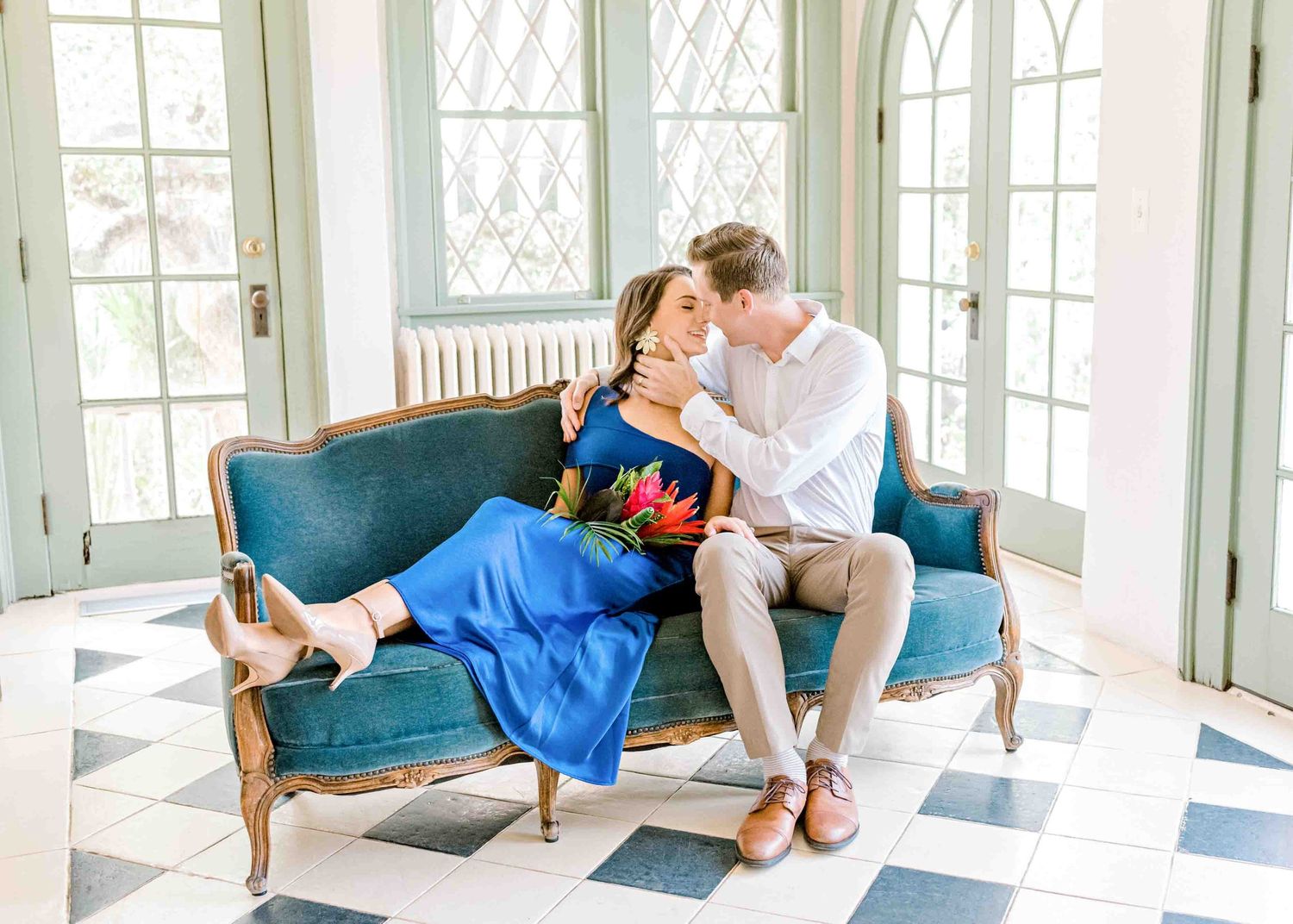 A couple shares a tender moment on a vintage blue sofa in a sunlit room with checkered floors.