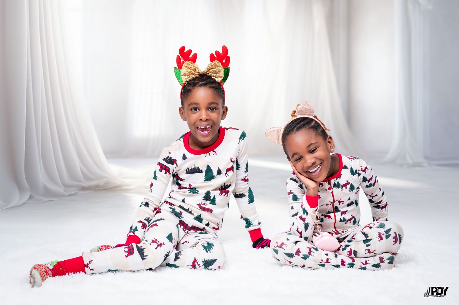 Siblings in festive white Christmas pajamas pose together wearing reindeer antler accessories.