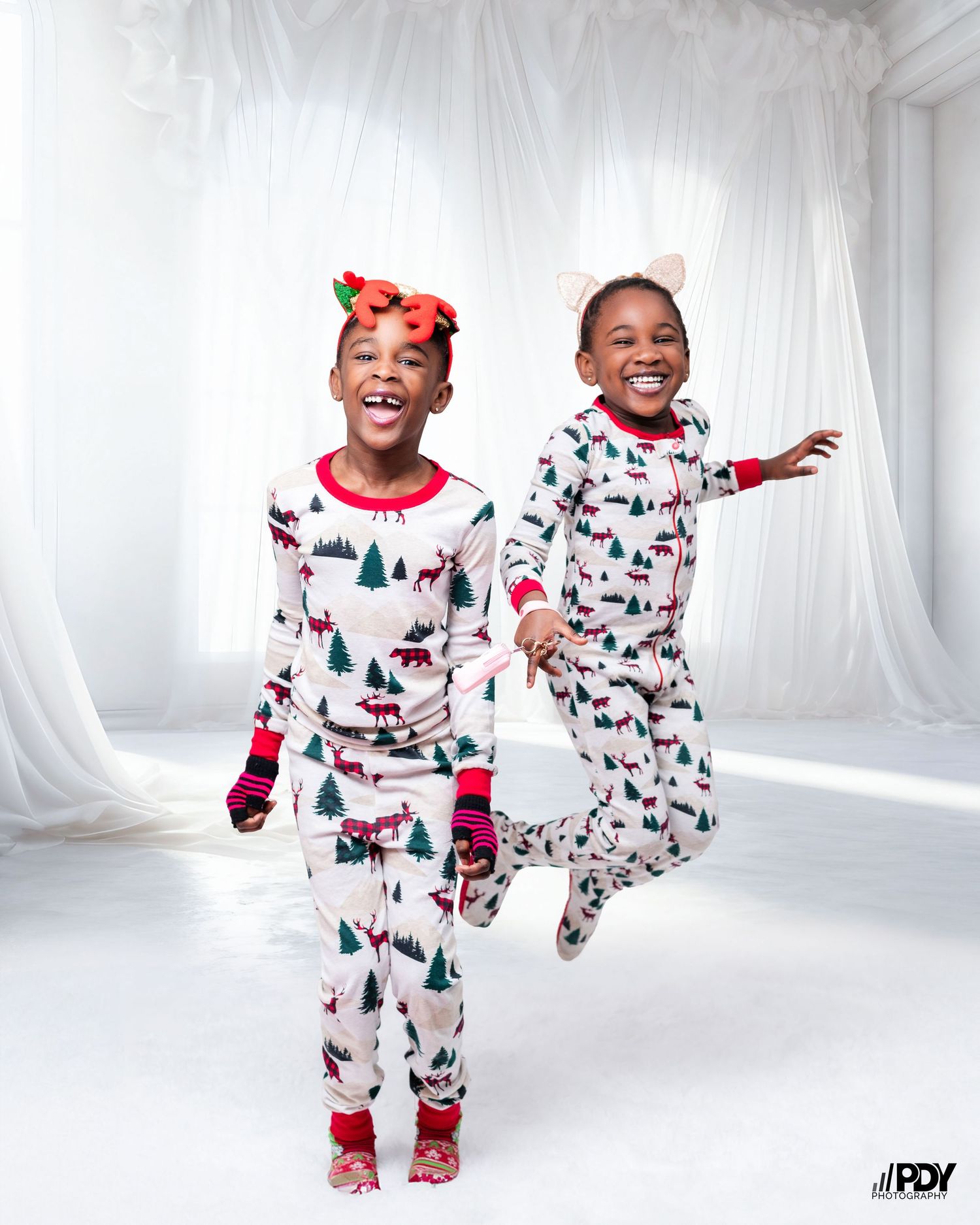 Two children in matching Christmas pajamas jumping joyfully against white backdrop.