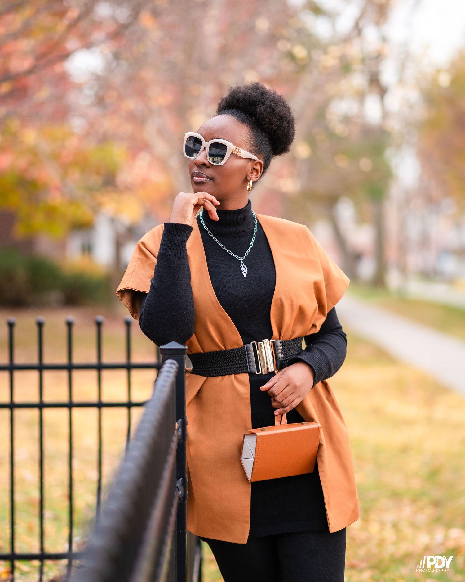 Stylish fall fashion look featuring an orange blazer, black top and sunglasses against autumn foliage.