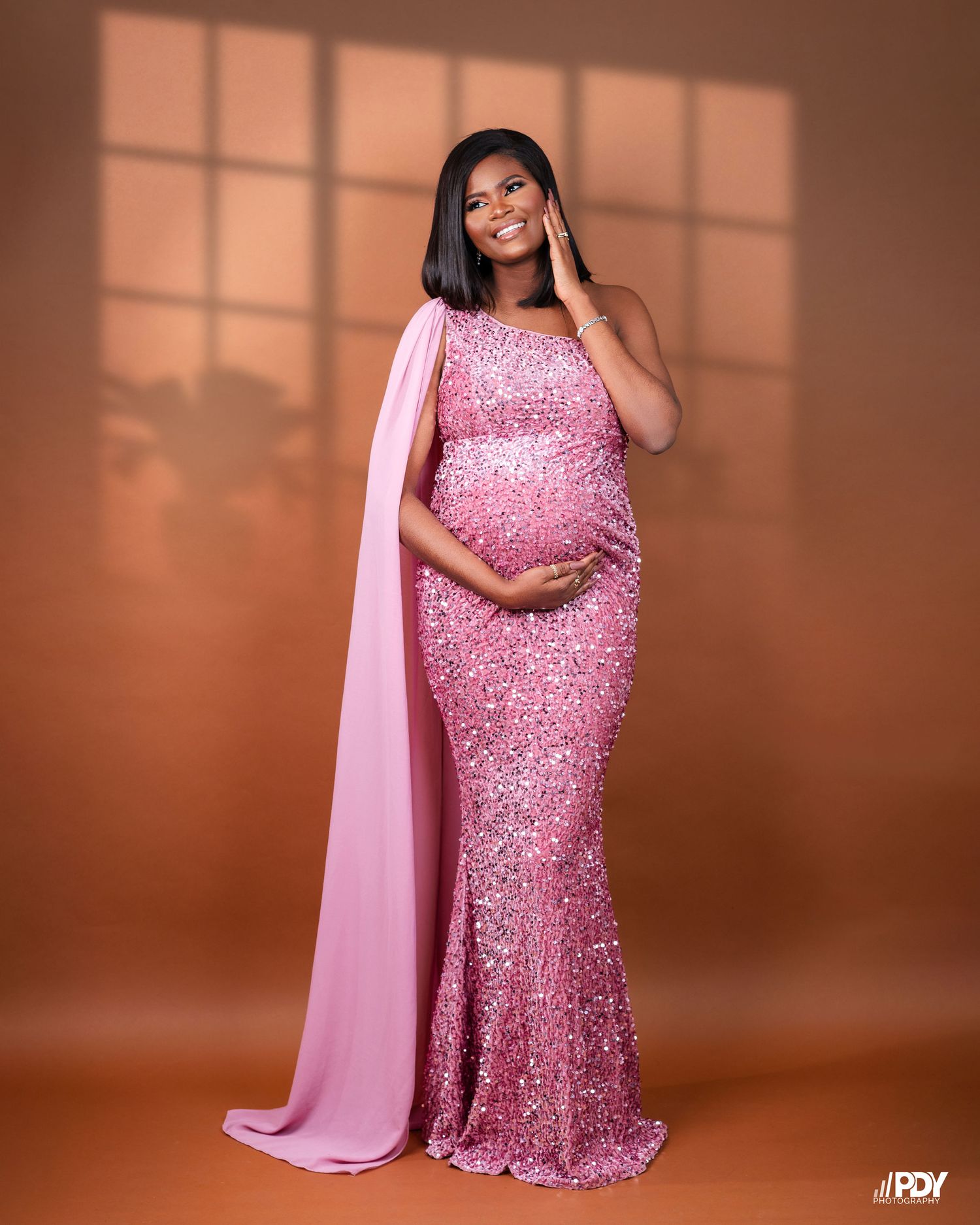 A stunning maternity photoshoot featuring a sparkly pink sequined gown and flowing cape against a warm brown backdrop.