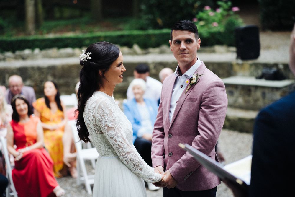 Outdoor wedding ceremony with couple in white dress and pink jacket exchanging vows.