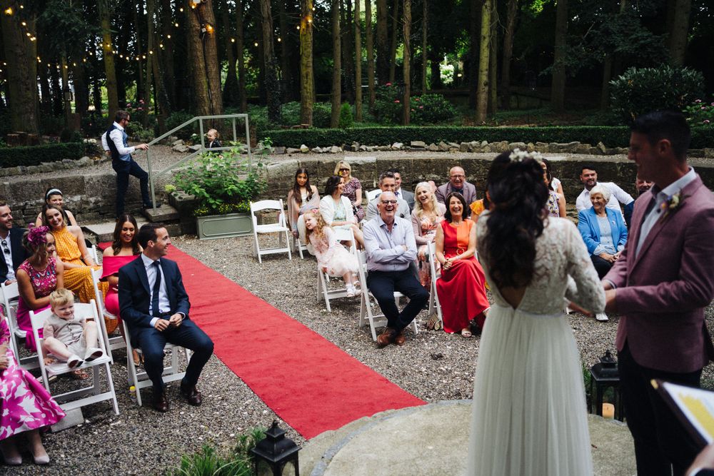 Outdoor wedding ceremony with guests seated on white chairs along a red carpet aisle in a wooded setting.