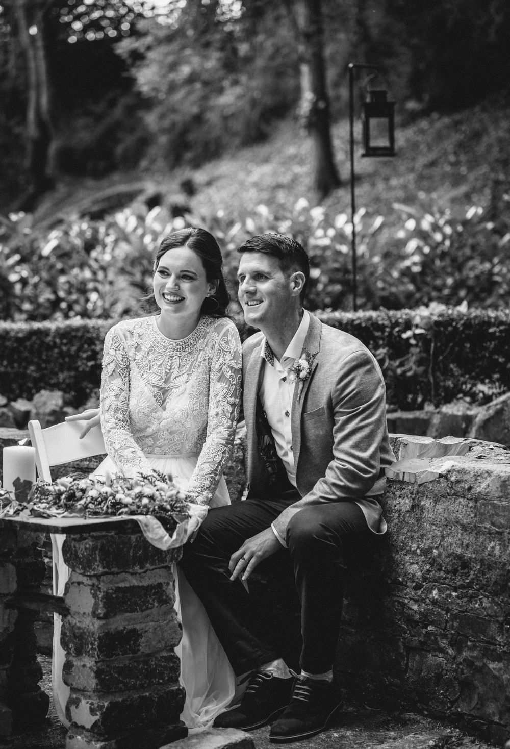 A couple sits together on a brick wall during their wedding day in a garden setting.