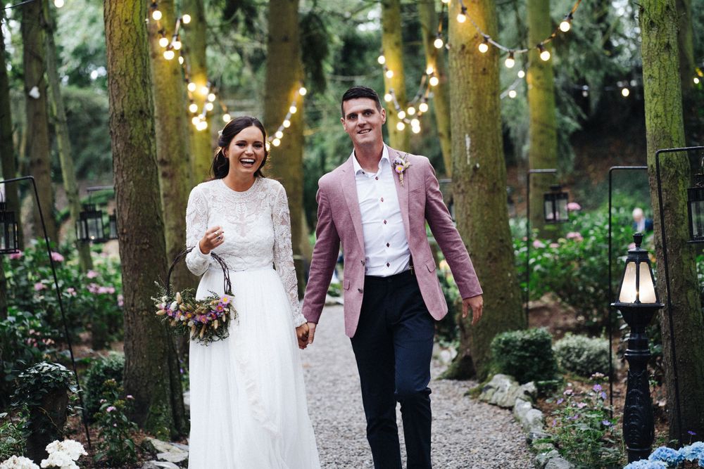 A wedding couple walks hand in hand down a tree-lined path decorated with string lights.