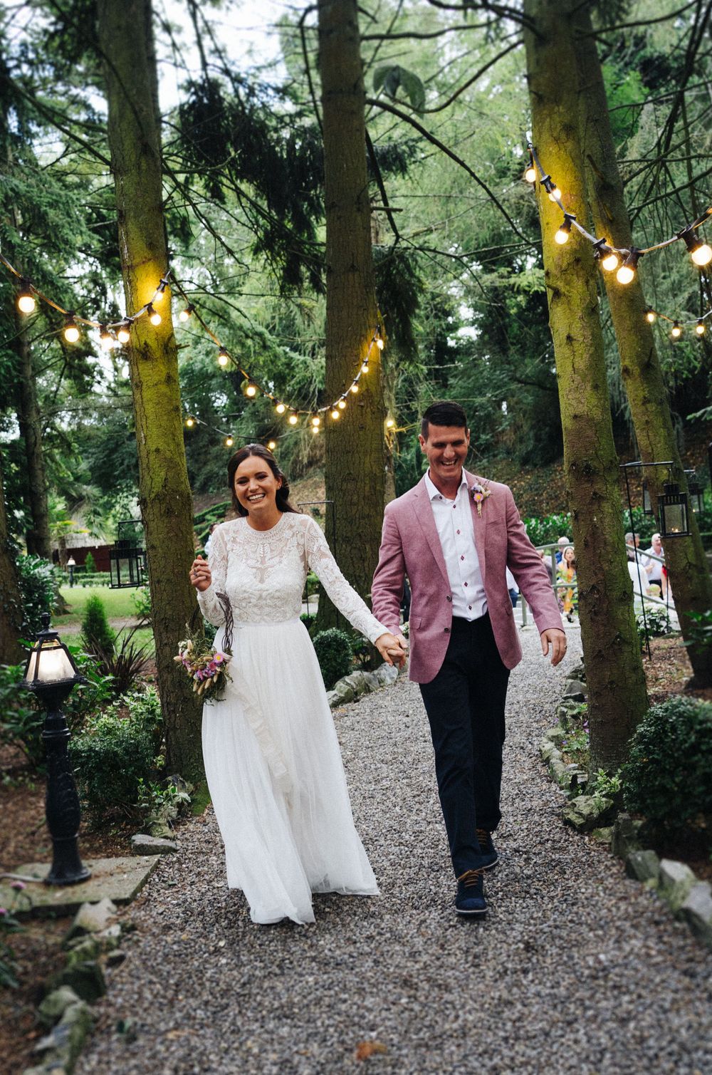 Newlyweds stroll together on a gravel path under trees decorated with twinkling lights.