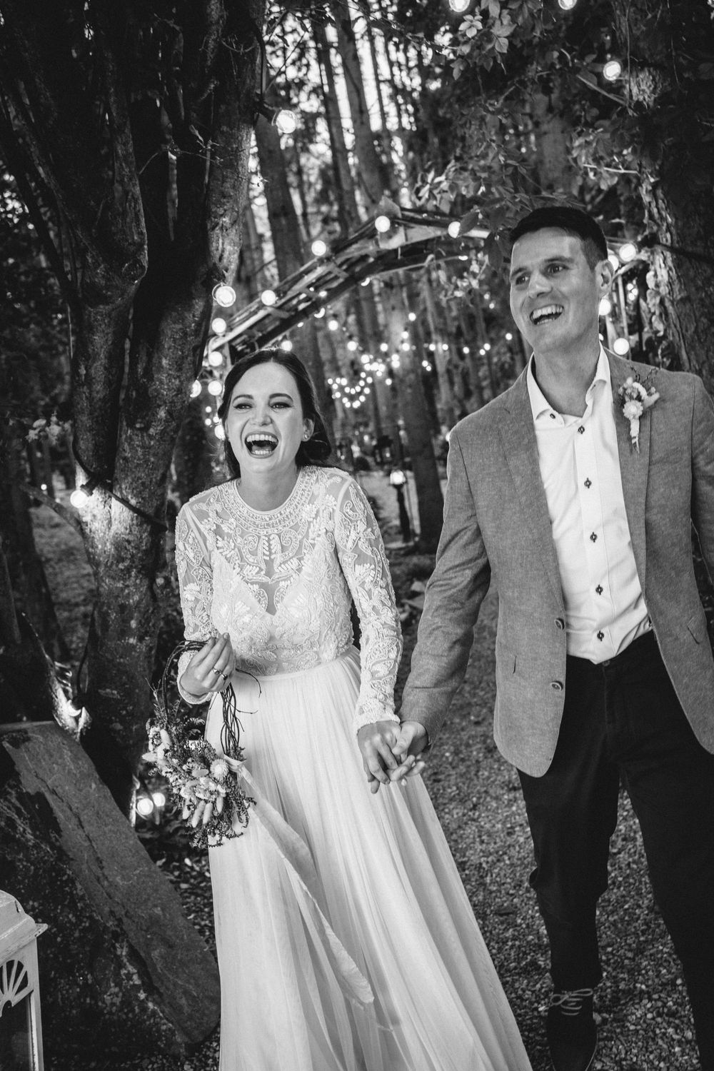 A couple shares a laugh while walking through a magical forest setting with string lights.
