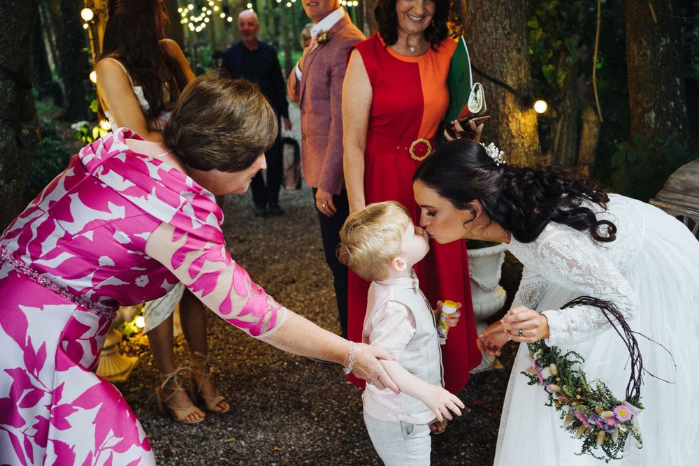 Wedding celebration outdoors with guests interacting near festive lighting and greenery.
