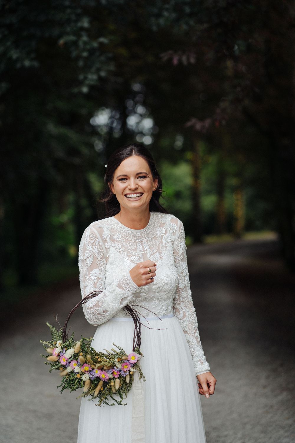 A person holding a floral bouquet stands on a wooded path wearing a long-sleeved lace wedding dress.