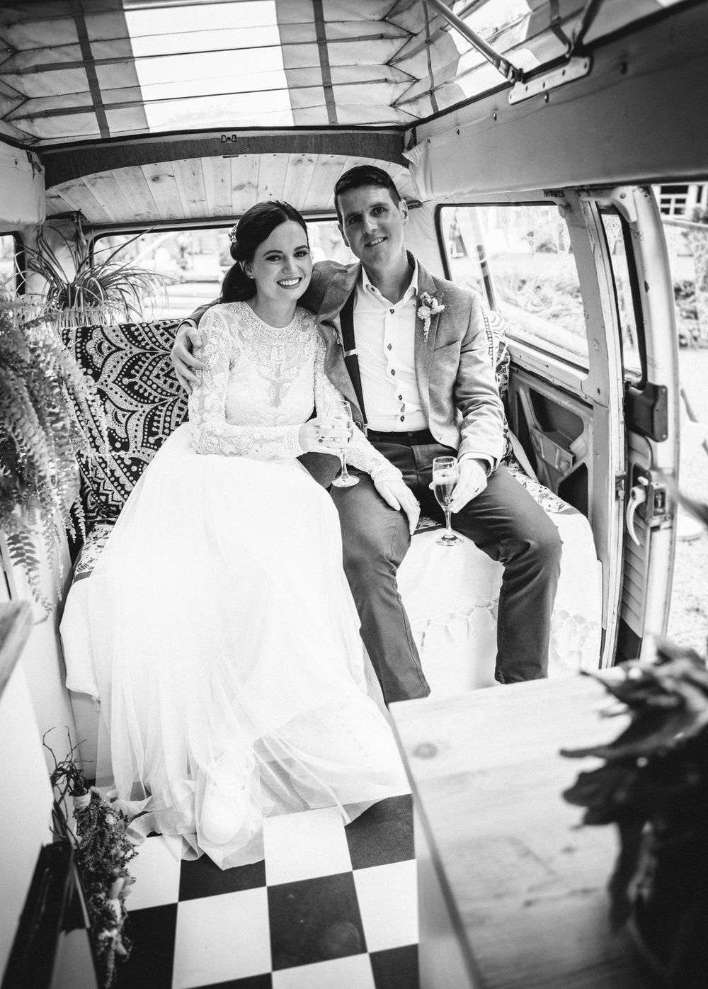 A vintage-style wedding photo showing a couple sitting in a retro van with checkered flooring and bohemian decor.