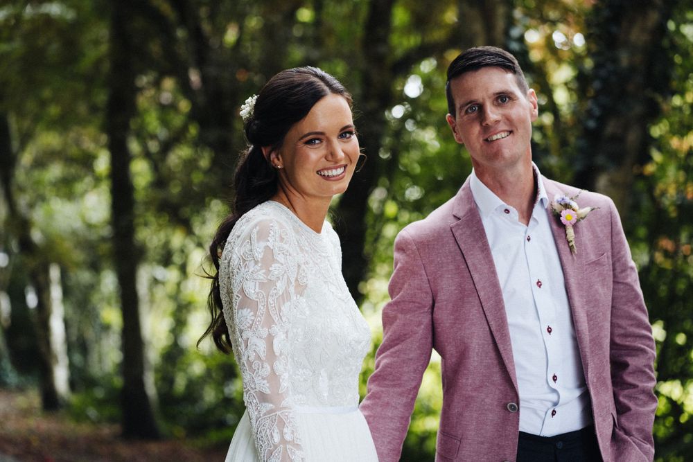 A romantic wedding couple posing in a sunlit forest wearing a lace gown and pink blazer.